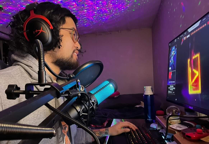 Cam Jr. Sengsuriya, a young man who has curly hair and wears orange glasses, streams a video game.