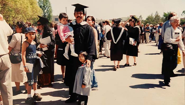 Mauricio dressed in a black grad cap and gown holding 1-year-old Fatima and surrounded but his other kids Ivonne, Karen, and Bonnie in the middle of parking lot filled with other people and grads.