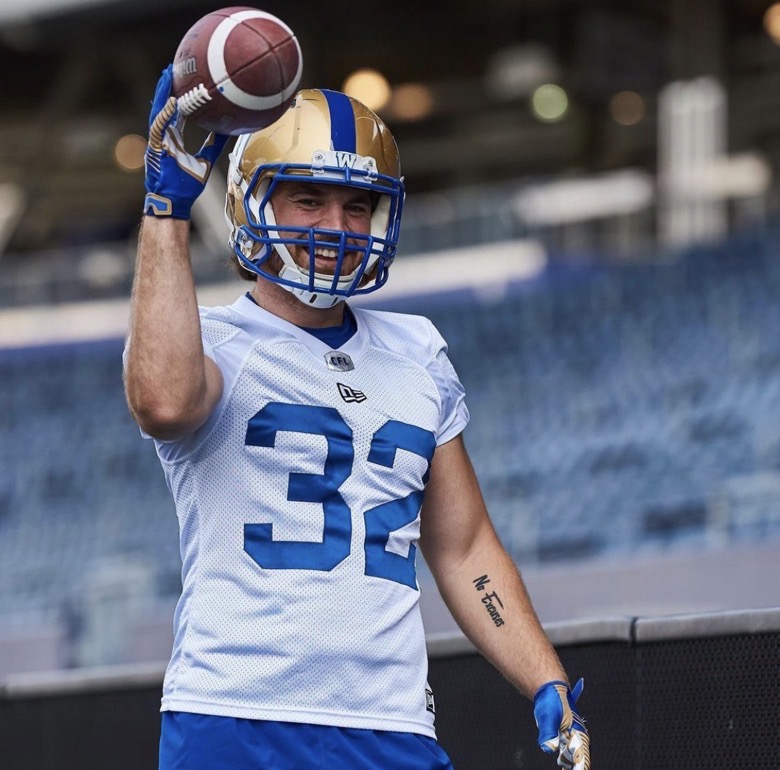 A football player, wearing a white jersey and holding a football in his right hand, smiles for the camera.