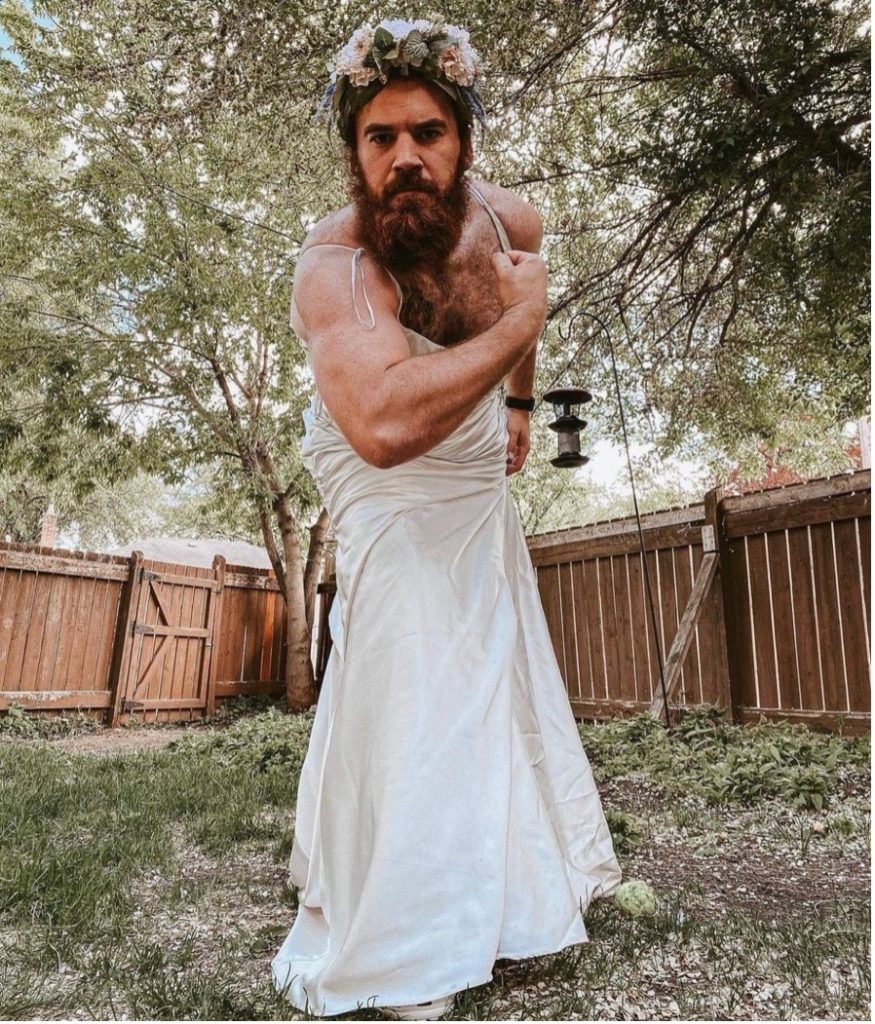 A muscular man with a full beard wears a white wedding dress as he flexes for the camera.