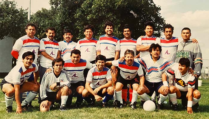 A group of middle aged latino men at a park wearing soccer uniforms,  arranged for a picture with nine in back row and seven in the front.