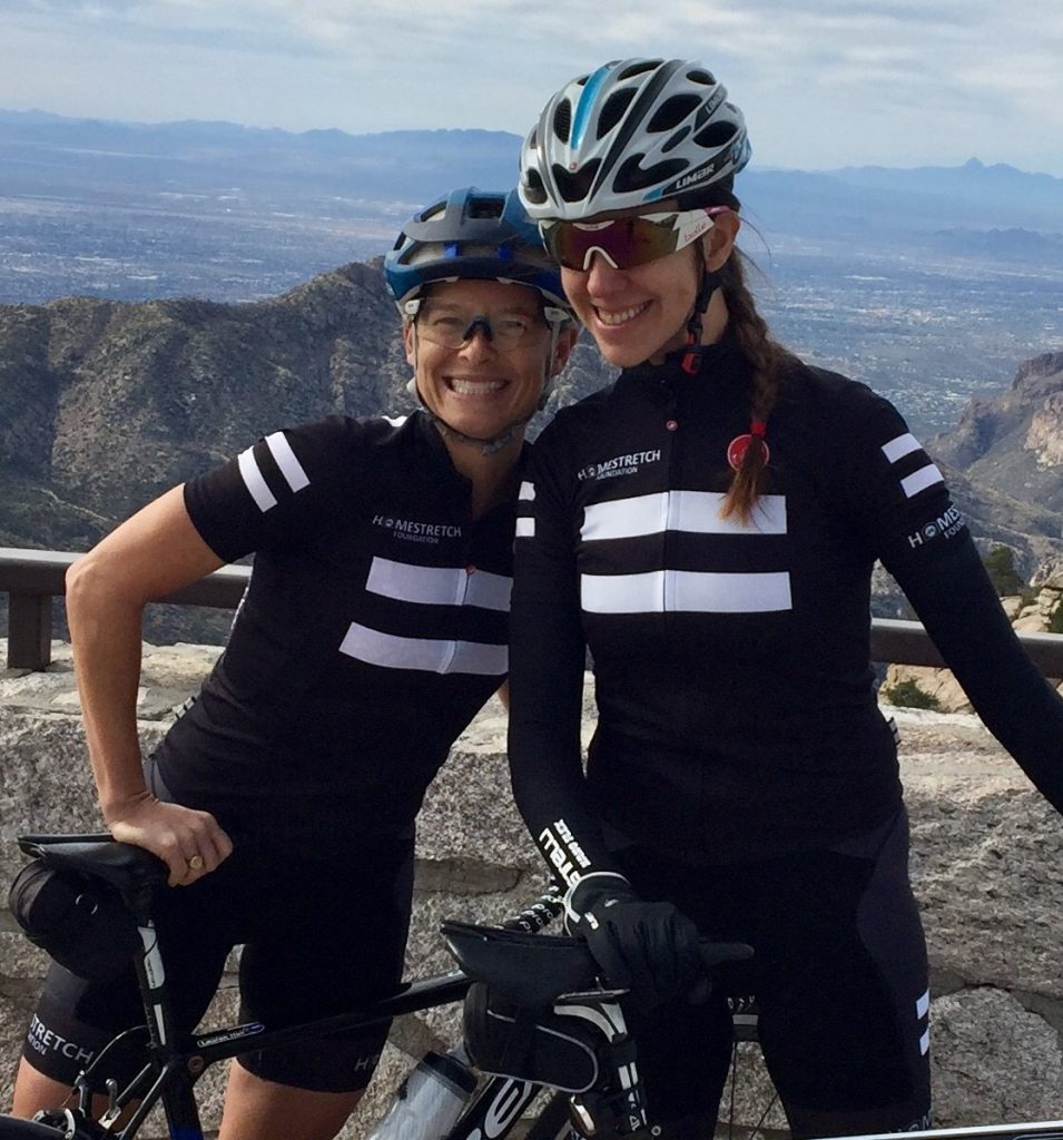 Two women in Homestretch cycling gear pose with their bikes in front of a mountain vista.
