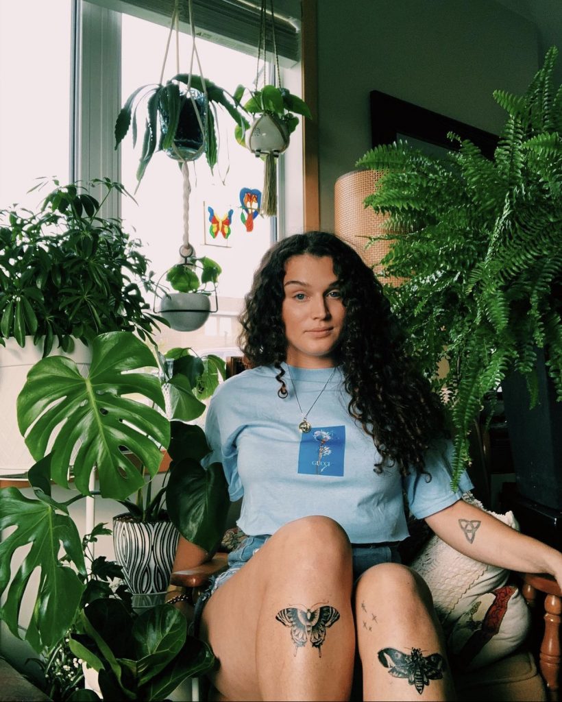 Sam Toews sits in a chair surrounded by houseplants, showing various tattoos on her arms and legs.