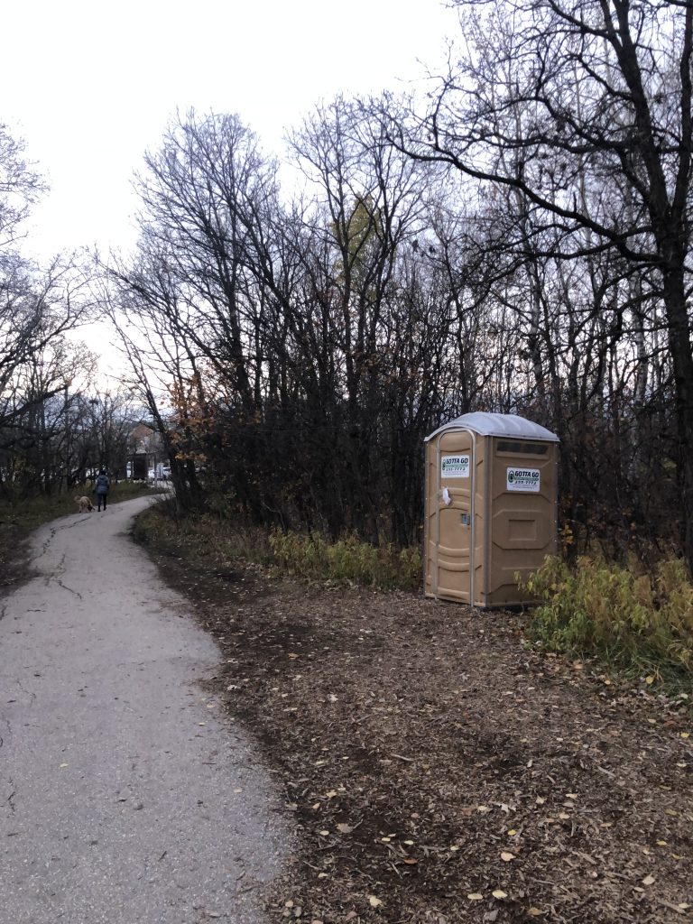 Image of a portable toilet at the entrance of the Assiniboine forest, on the only fully paved trail.