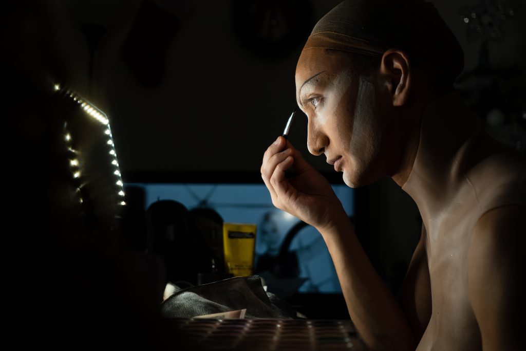 An image of Lady Fortuna applying her eyebrows as she carefully looks at herself through a vanity mirror. 